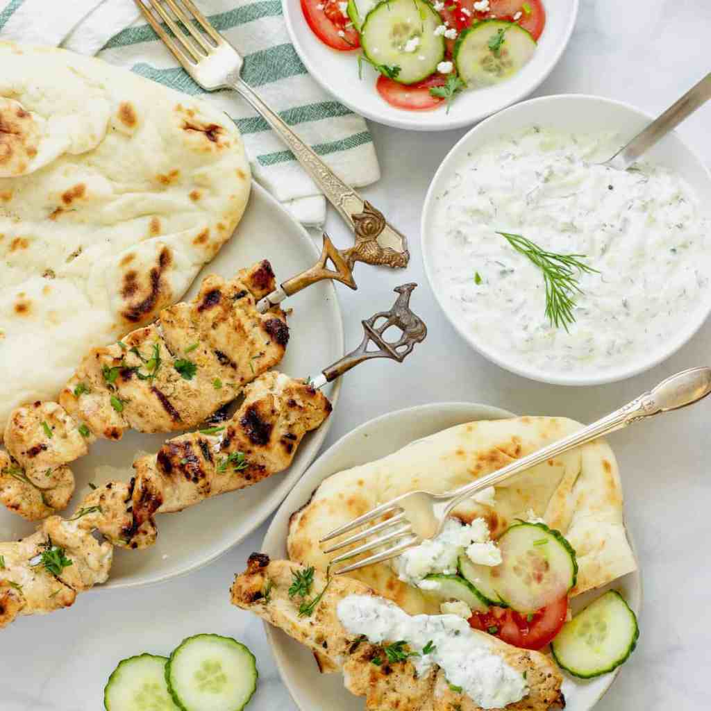 A plate of grilled chicken skewers garnished with herbs, accompanied by naan bread, a fresh cucumber and tomato salad, and a creamy yogurt sauce.