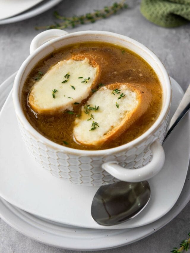 A bowl of French onion soup topped with two slices of melted cheese on toasted bread, garnished with fresh thyme, sitting on a gray surface.