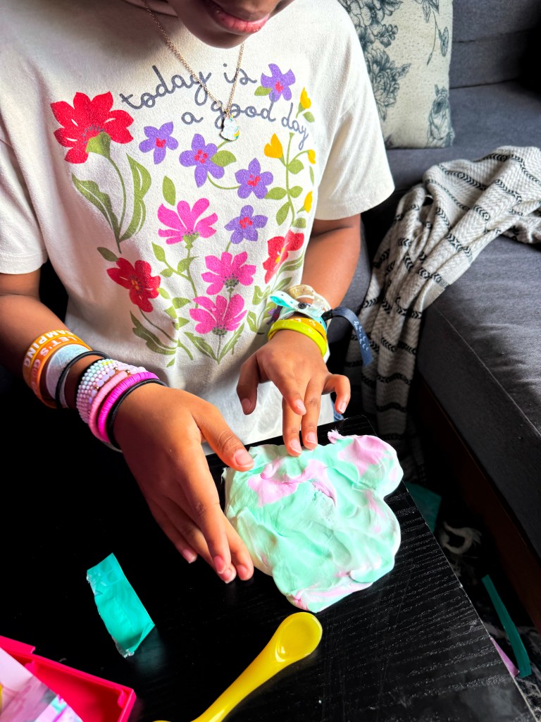 A child wearing a floral t-shirt is playing with colorful dough on a table, surrounded by crafting materials.