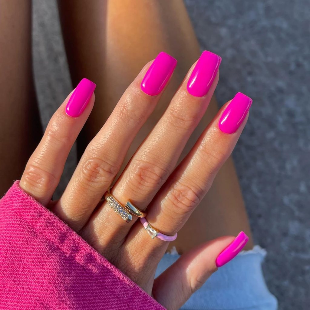A close-up of a hand with long, bright pink nails adorned with rings, set against a textured background.