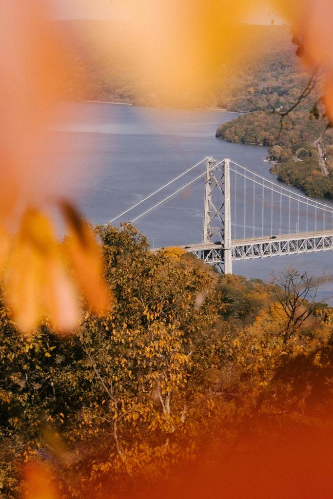 A scenic view of a bridge over a calm river, framed by vibrant autumn leaves in shades of orange and yellow.