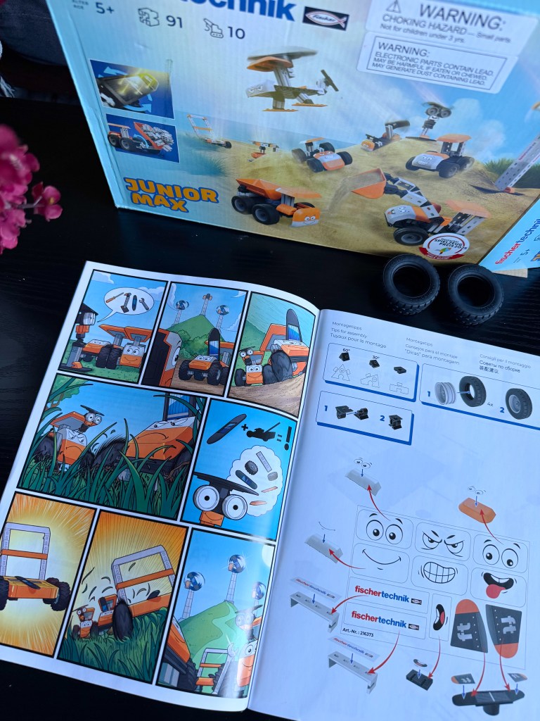 Close-up of a fischertechnik Junior Max building kit box featuring colorful toy designs and a warning label, alongside an open instruction booklet with comic-style illustrations and assembly instructions.