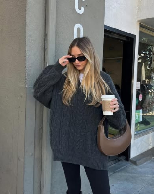 A person wearing a cozy oversized gray cable-knit sweater, black leggings, and sunglasses, holding a coffee cup while standing near a storefront.