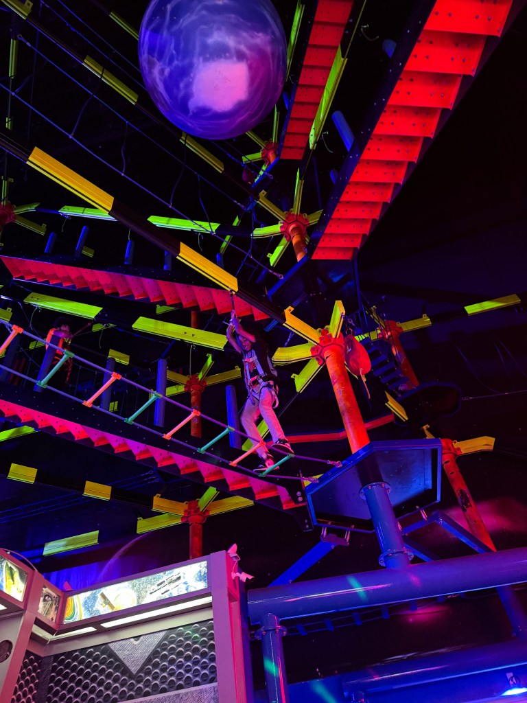 A person navigating an indoor ropes course suspended from a colorful framework with bright neon elements and a large planet-shaped decoration overhead.