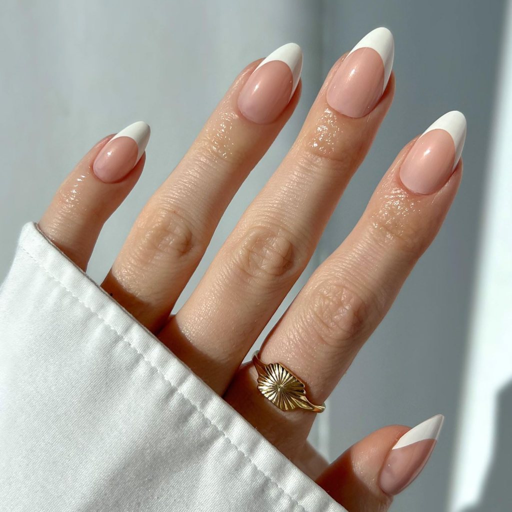 A close-up of a hand with well-manicured nails featuring a nude base color and white French tips, wearing a gold ring with a leaf design, resting on a white sleeve.