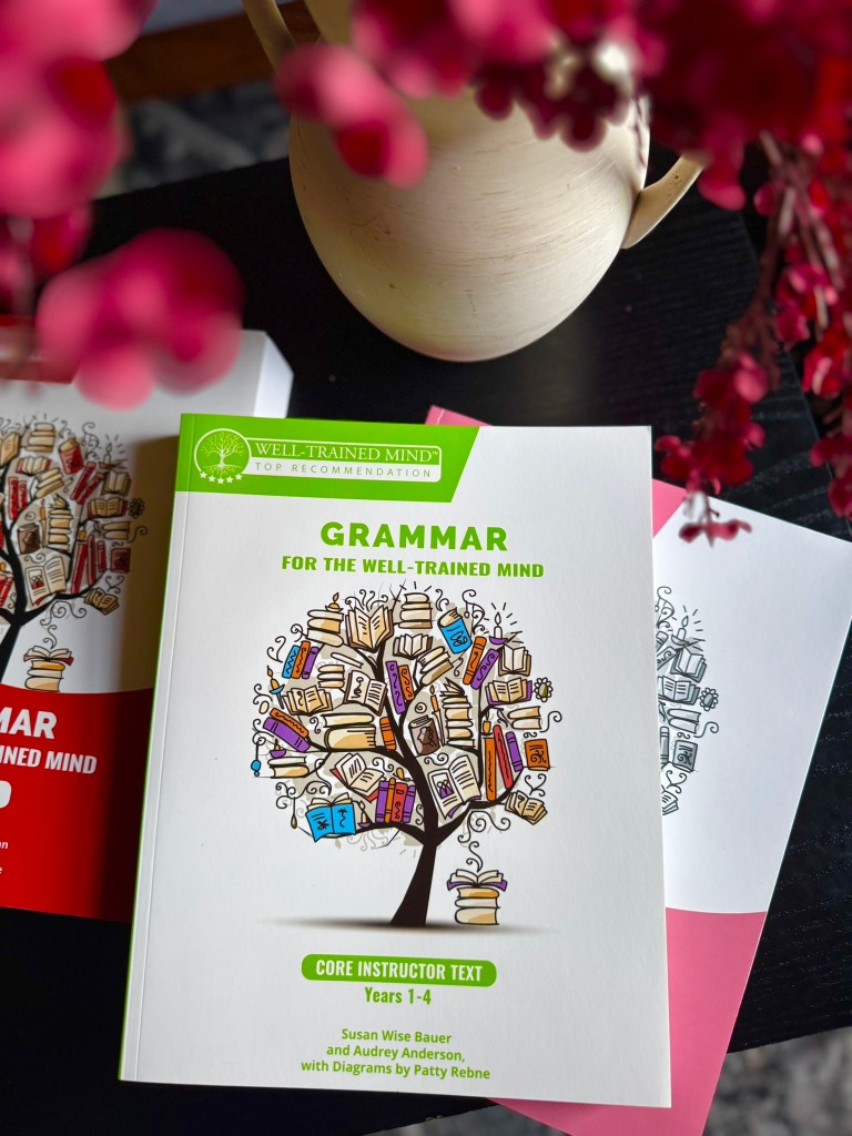 A close-up of the book 'Grammar for the Well-Trained Mind', featuring a colorful tree design with books as leaves, placed on a table with decorative flowers in the background.