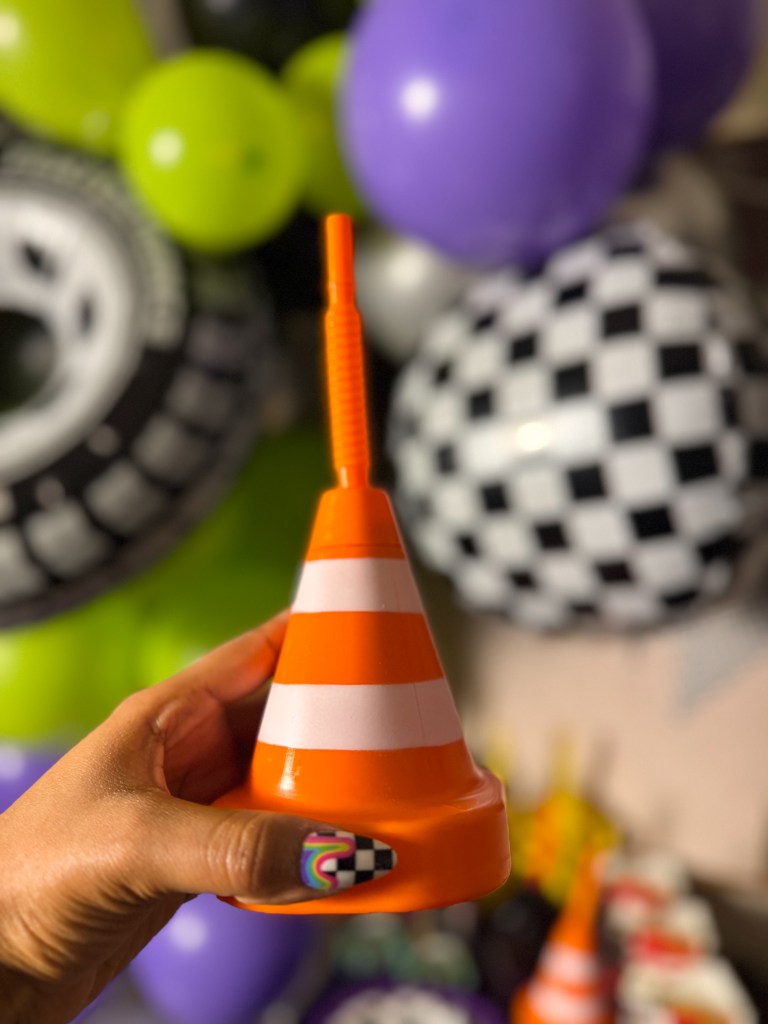 Hand holding an orange traffic cone decoration cup in front of Monster Jam birthday party decorations with balloons and racing theme.