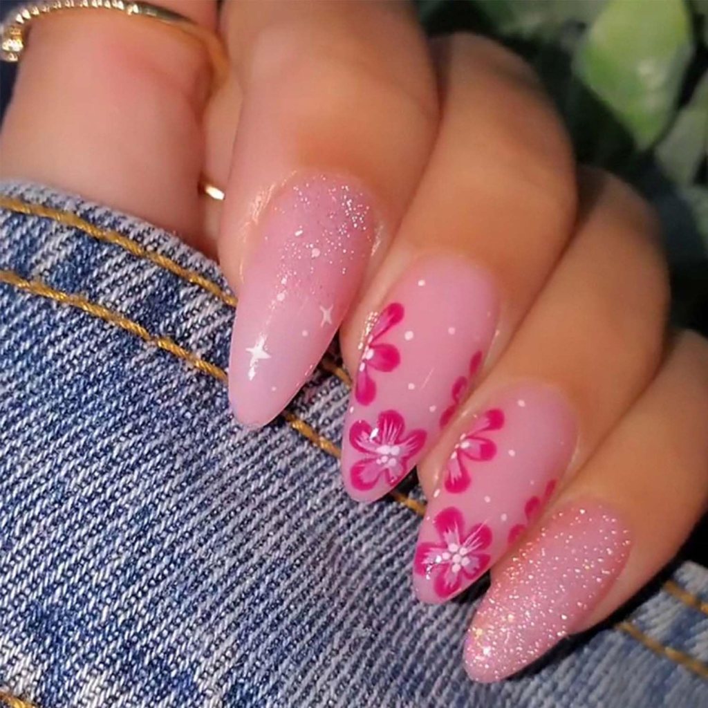 Close-up of a hand with long, almond-shaped nails painted in a soft pink color, featuring glitter accents and intricate floral designs in shades of pink and white, resting on a denim surface.