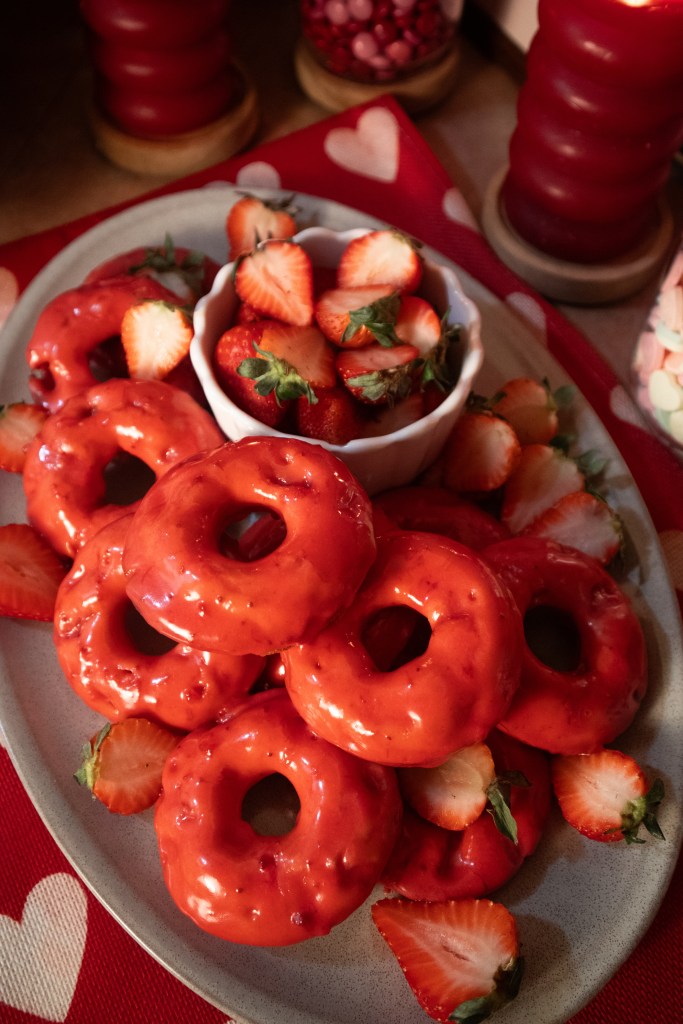 A plate of red-glazed vegan strawberry donuts arranged with fresh strawberries on a Valentine's Day themed table.