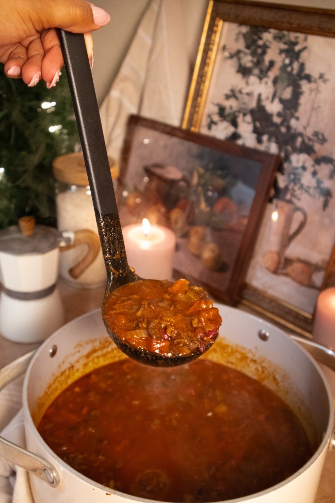 A hand holding a ladle filled with a hearty stew, with a pot of stew in the foreground and decorative items in the background, including candles and framed pictures.