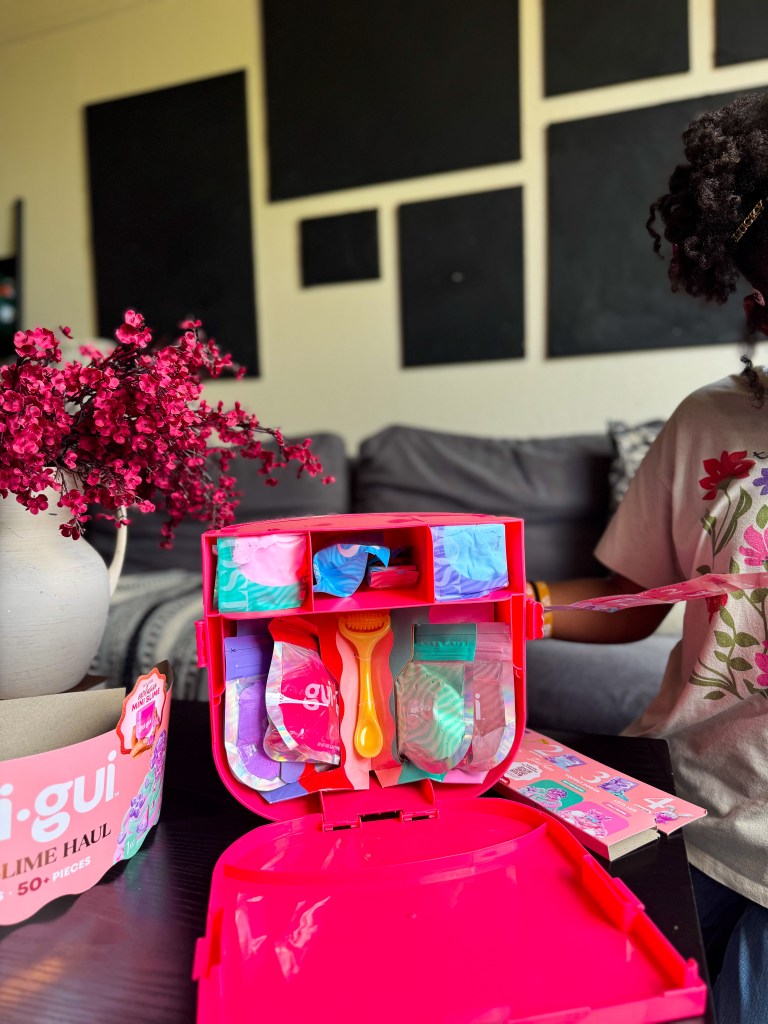 A brightly colored pink slime kit open on a table, featuring various slime components and tools, with a child partially visible in the background and a bouquet of pink flowers in the foreground.