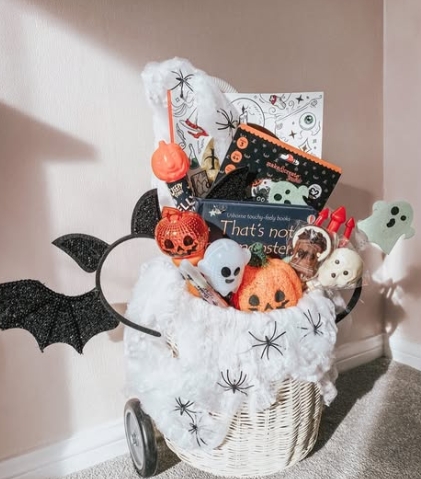 A Halloween-themed wicker basket filled with plush toys, decorations, and books, including a black cat and ghost items, topped with a cozy white blanket and bat accents.