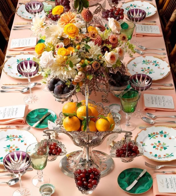 A beautifully arranged Thanksgiving table featuring a floral centerpiece with orange and yellow flowers, surrounded by elegant dinnerware, colorful glassware, and assorted fruits including oranges, plums, and grapes.