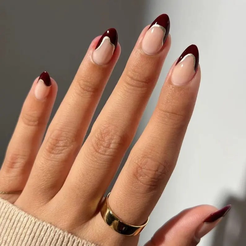 A close-up of a hand displaying manicured nails with a burgundy tip and a subtle silver line, wearing a gold ring.