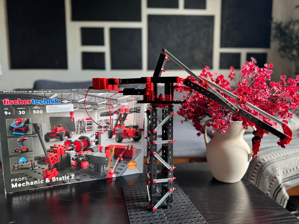A Fischer Technik STEM Kit displayed on a table, featuring a model crane made from red and black building pieces. The box for the kit is visible in the background, showcasing various models and components.