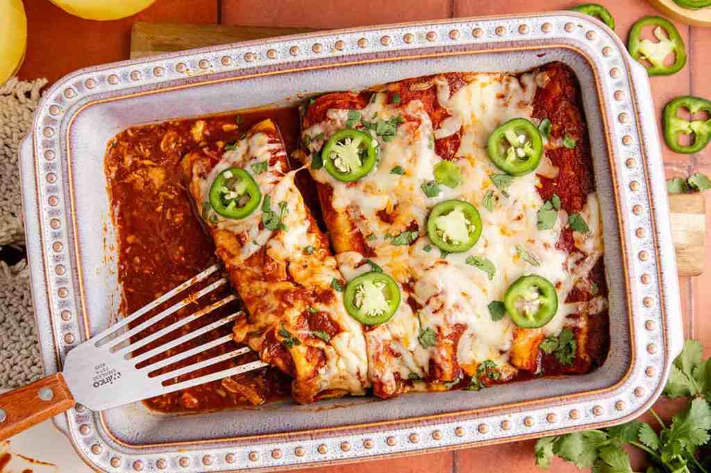 A square baking dish filled with cheesy chicken enchiladas topped with sliced jalapeños and fresh cilantro, served with a spatula on the side.