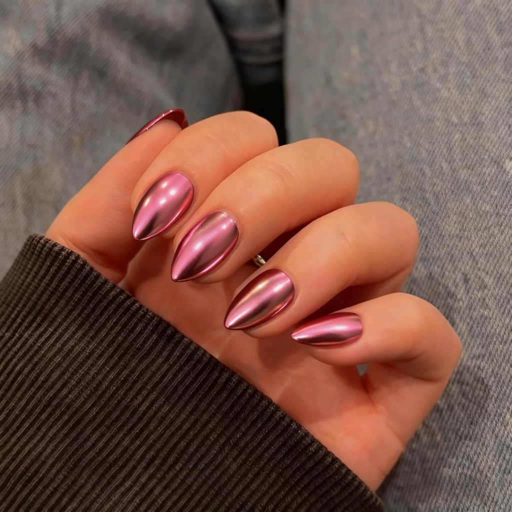 Close-up of a hand with long, pointed nails painted in a shiny metallic pink color, resting on a textured surface.