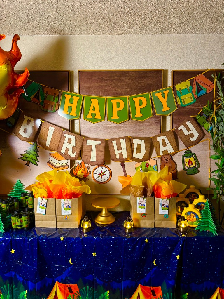 Camping birthday party backdrop with happy birthday banner, favor bags, and outdoor adventure decorations.