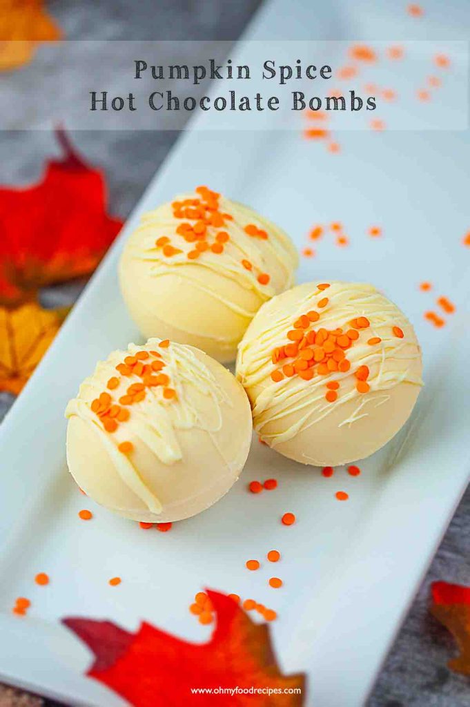 Three pumpkin spice hot chocolate bombs on a white plate, decorated with orange and yellow sprinkles, surrounded by autumn leaves.