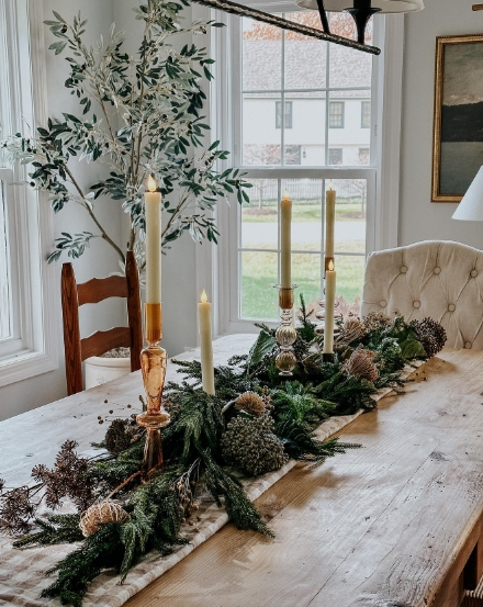 A cozy fall dining table featuring a centerpiece of greenery, candles in various heights, and natural decorations like pinecones, set against a bright window backdrop.
