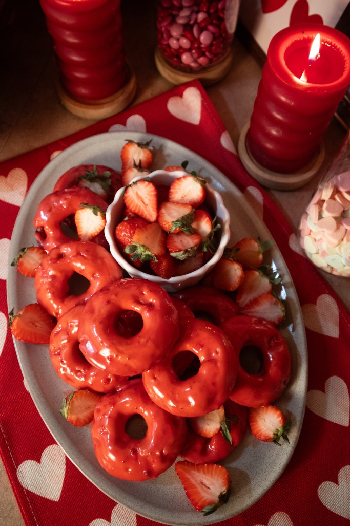 A plate of red-glazed donuts surrounded by sliced strawberries, with candles and decorative jars in the background.