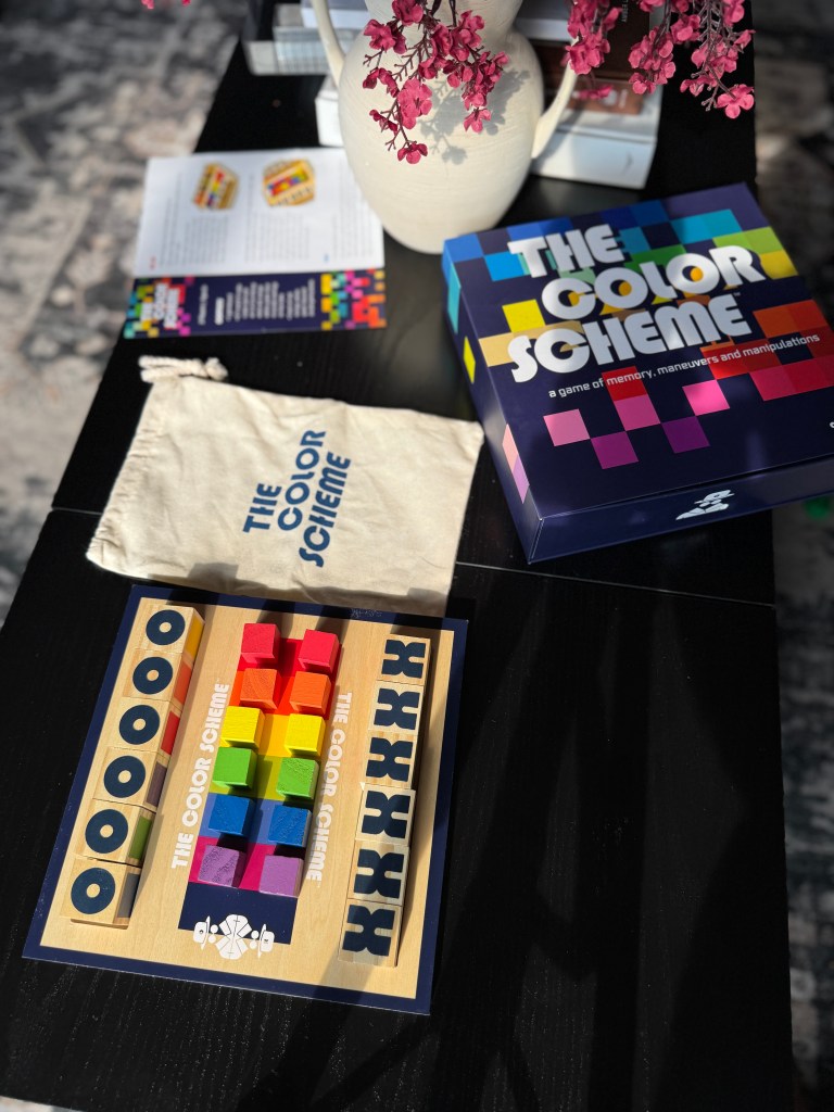 A colorful board game called 'The Color Scheme' displayed on a black table, showing vibrant game pieces and a soft canvas pouch.
