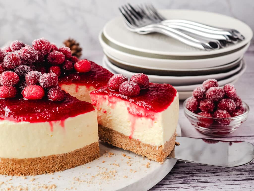 A delicious cheesecake topped with a red fruit sauce and fresh sugared cranberries, served on a white marble board with a small bowl of extra cranberries and a set of plates in the background.