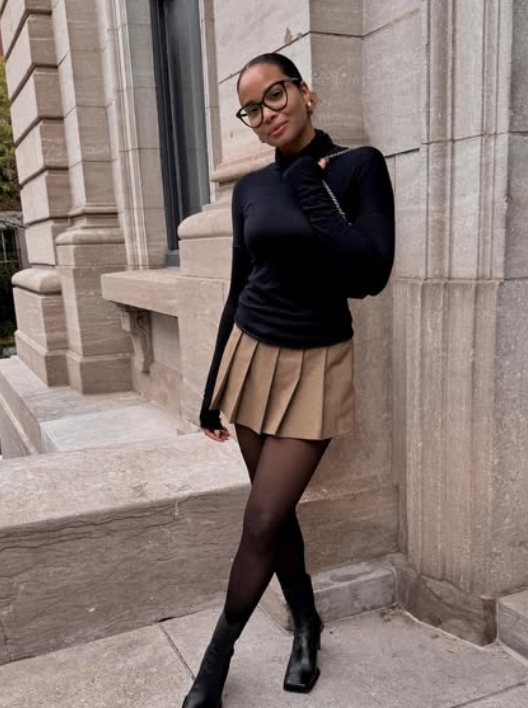 A young woman poses confidently in a fitted black turtleneck and a pleated khaki mini skirt, complemented by sheer black tights and ankle boots. She wears glasses and stands against a stone backdrop.