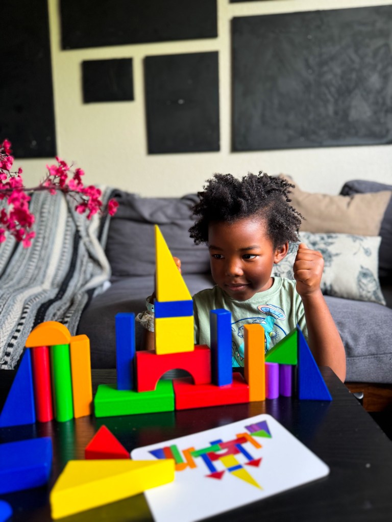 A young child with curly hair is sitting in front of a colorful arrangement of geometric building blocks, showing an expression of concentration. The setting is a cozy living space with a couch and dark wall panels in the background.