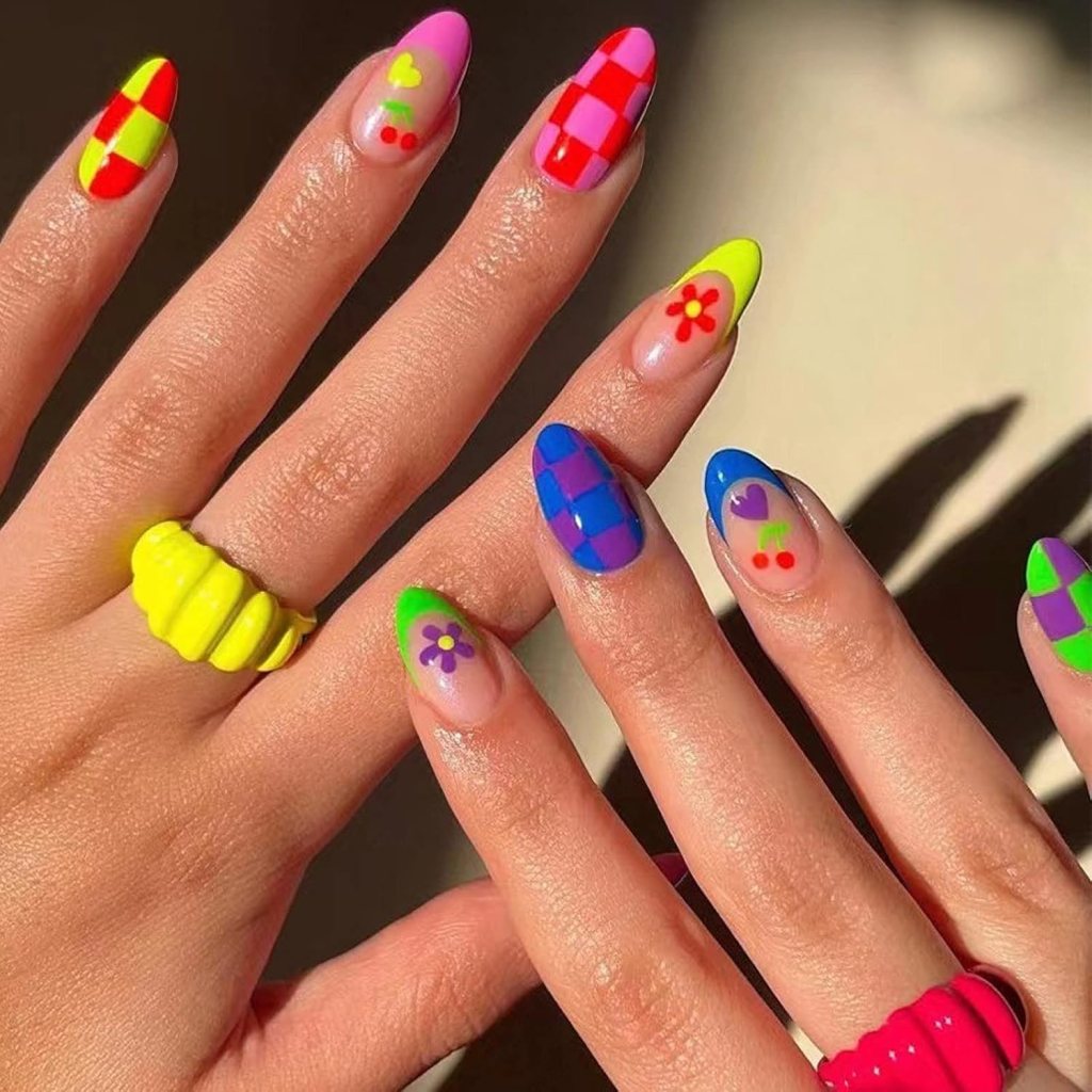 Close-up of two hands with brightly colored nail art featuring various patterns, including hearts, checks, flowers, and fruit designs. The nails are shades of pink, red, yellow, blue, and green. One hand wears a bright yellow ribbed ring and the other a pink ring.