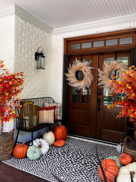 A cozy fall-inspired porch featuring colorful pumpkins, vibrant foliage, and decorative wreaths adorning the double wooden doors, creating a warm and inviting entrance.