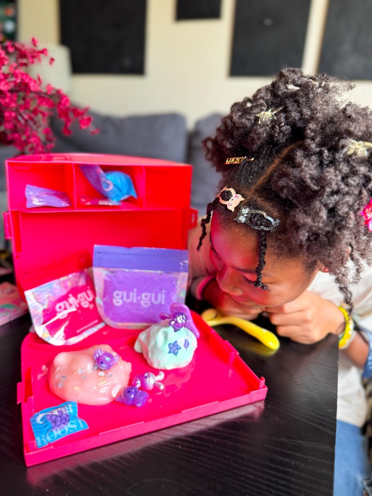 A young child looking curiously at colorful slime in a bright pink craft box, surrounded by playful decorations and materials.