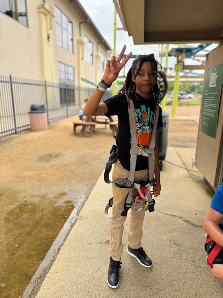 A young boy wearing a safety harness and a black graphic t-shirt, giving a peace sign outdoors near a building.
