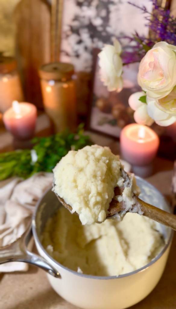 A wooden spoon holding a serving of creamy mashed potatoes above a pot of mashed potatoes, with candles and flowers in the background.