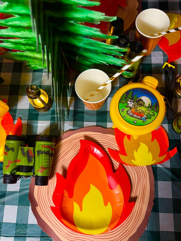 Camping birthday party place setting with fire napkins, wooden cups, toy binoculars, and yellow canteens.