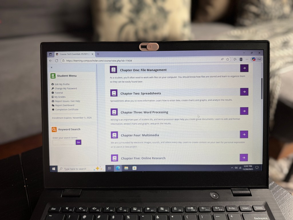 A laptop screen displaying the student dashboard for CompuScholar's Tech Essentials course, featuring chapters on file management, spreadsheets, word processing, multimedia, and online research.