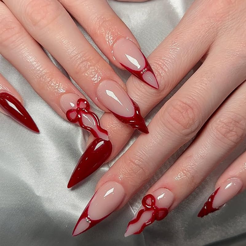 Close-up of hands with stylish nail art featuring long, pointed nails painted in a combination of nude and dark red, some adorned with decorative bows.