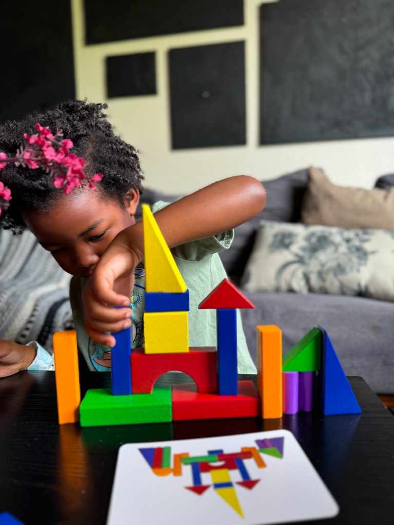 A young child with curly hair and flowers in their hair is building a colorful structure using various geometric blocks on a black table, with a design card visible in front.