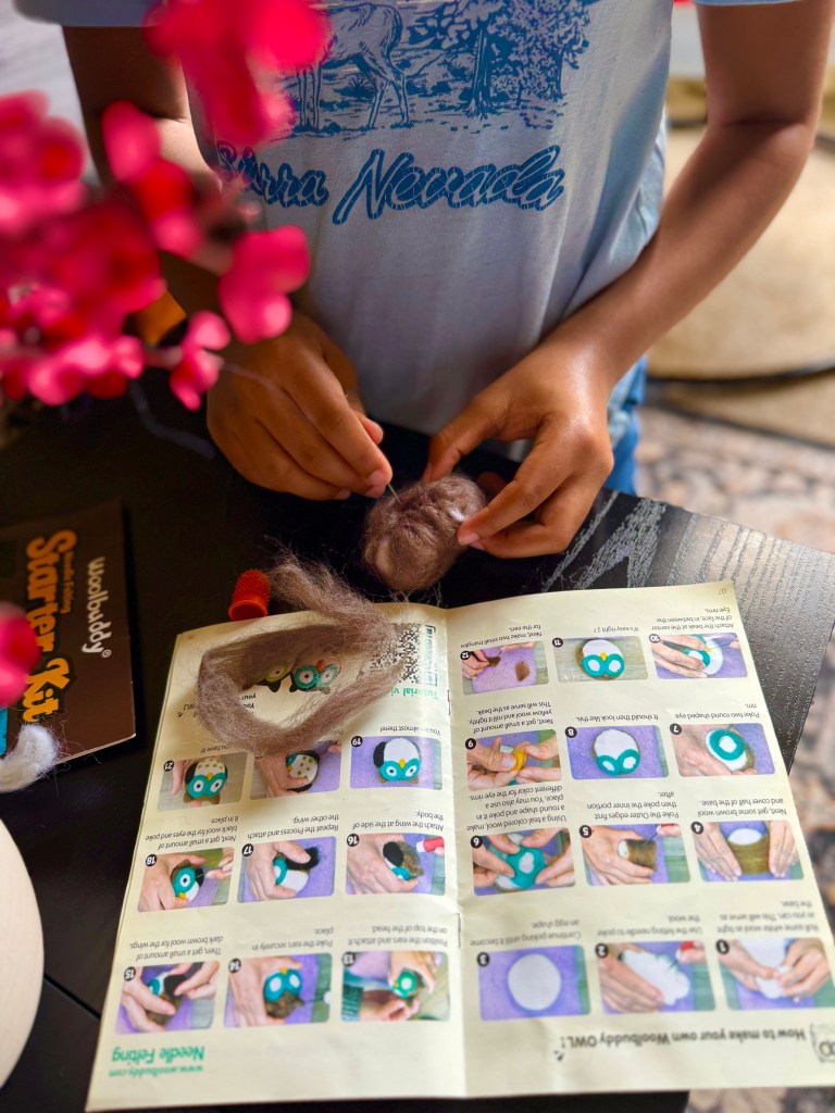 A child following a needle felting tutorial, holding wool and a felting needle, with an instructional booklet open beside them.