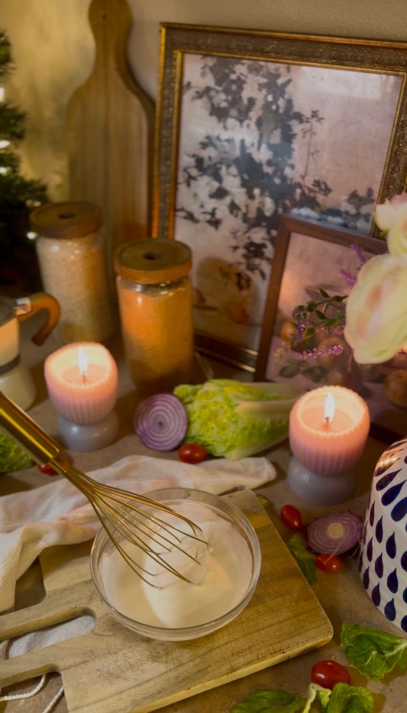 A kitchen scene featuring a whisking bowl of cream on a wooden cutting board, surrounded by fresh vegetables like lettuce, tomatoes, and red onions. Decorative candles provide soft lighting, and a vintage framed artwork is visible in the background.