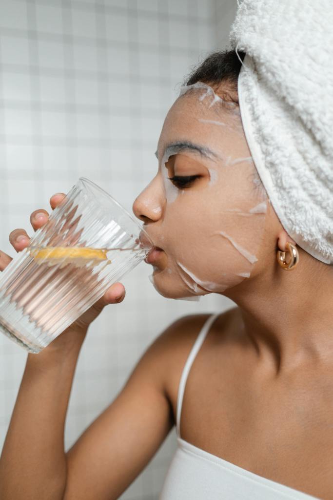 A person with a facial mask drinks lemon water from a glass, showcasing a moment of self-care and hydration.