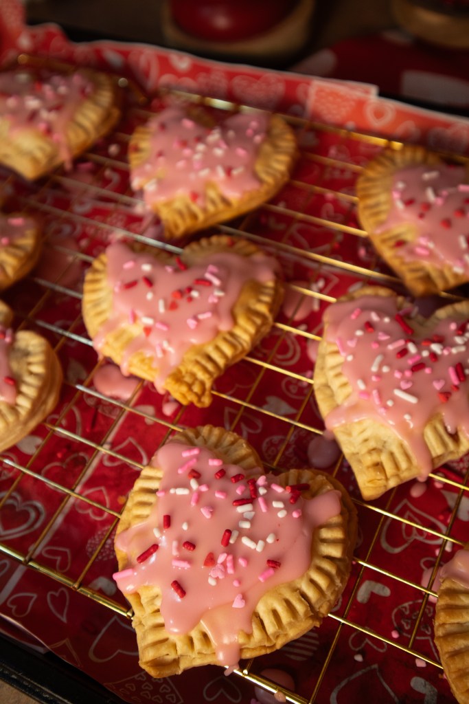 Freshly baked heart-shaped pop tarts decorated with pink icing and colorful sprinkles, displayed on a cooling rack.