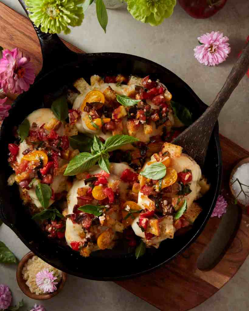 A cast-iron skillet filled with baked chicken topped with colorful diced tomatoes, herbs, and bread crumbs, garnished with fresh basil leaves and surrounded by flowers and a bowl of grated cheese.