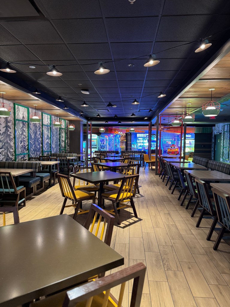 Interior of the dining area at Great Wolf Lodge, featuring wooden tables and chairs, a cozy atmosphere, and colorful décor, with visible arcade games in the background.