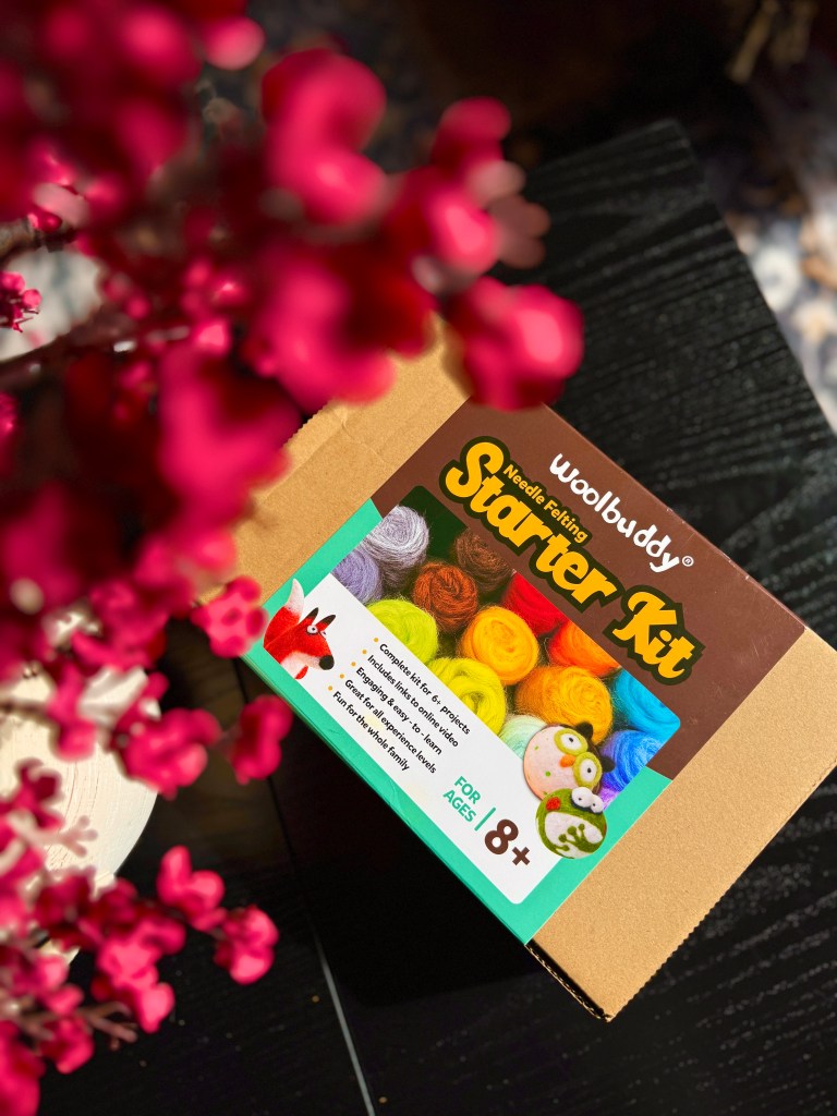An image of a Woolbuddy Needle Felting Starter Kit box, featuring vibrant colored wool roving and playful illustrations of a fox and an owl, surrounded by pink flowers on a dark surface.