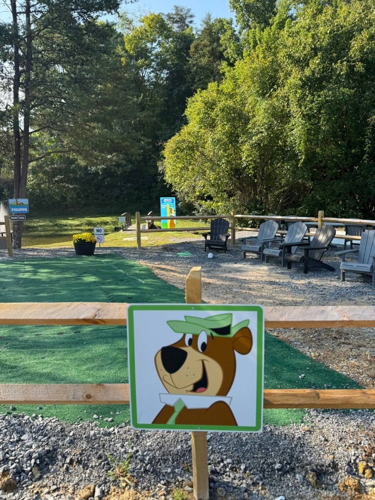 A playful cartoon bear character sign is displayed in a recreational area with seating and greenery in the background.