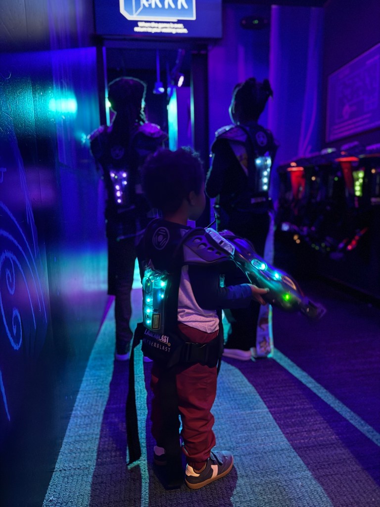 A young child in a laser tag arena, wearing a glowing vest and holding a laser gun, stands in a corridor with other players in the background.