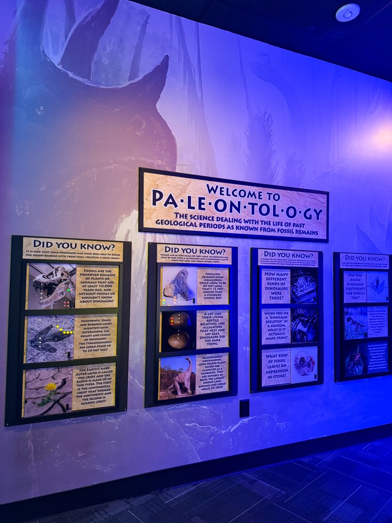 Exhibit entrance to a paleontology display featuring educational panels and fossil-related information, with a dinosaur mural in the background.