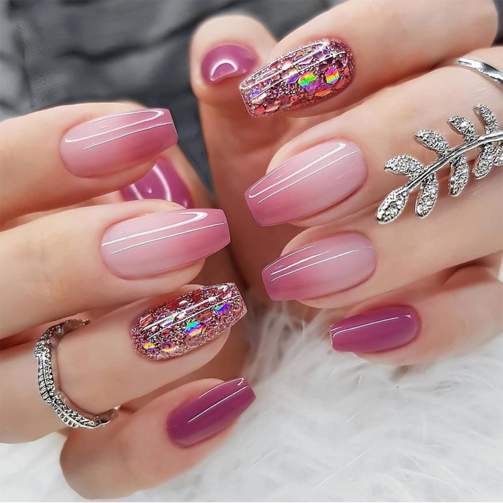 Close-up of manicured nails featuring an ombre design in pink shades and one accent nail with glitter and holographic details. A silver ring and a decorative leaf embellishment are visible.