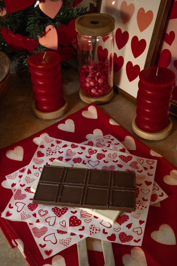 A festive display featuring dark chocolate on heart-themed paper, surrounded by red candles, a jar of candies, and decorations with hearts.